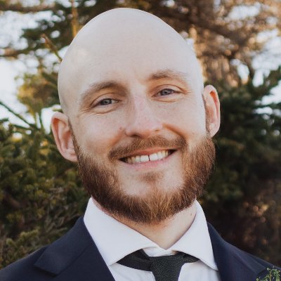 My professional headshot. A white man with a shaved head and a beard, wearing a suit and standing in front of a forested background.